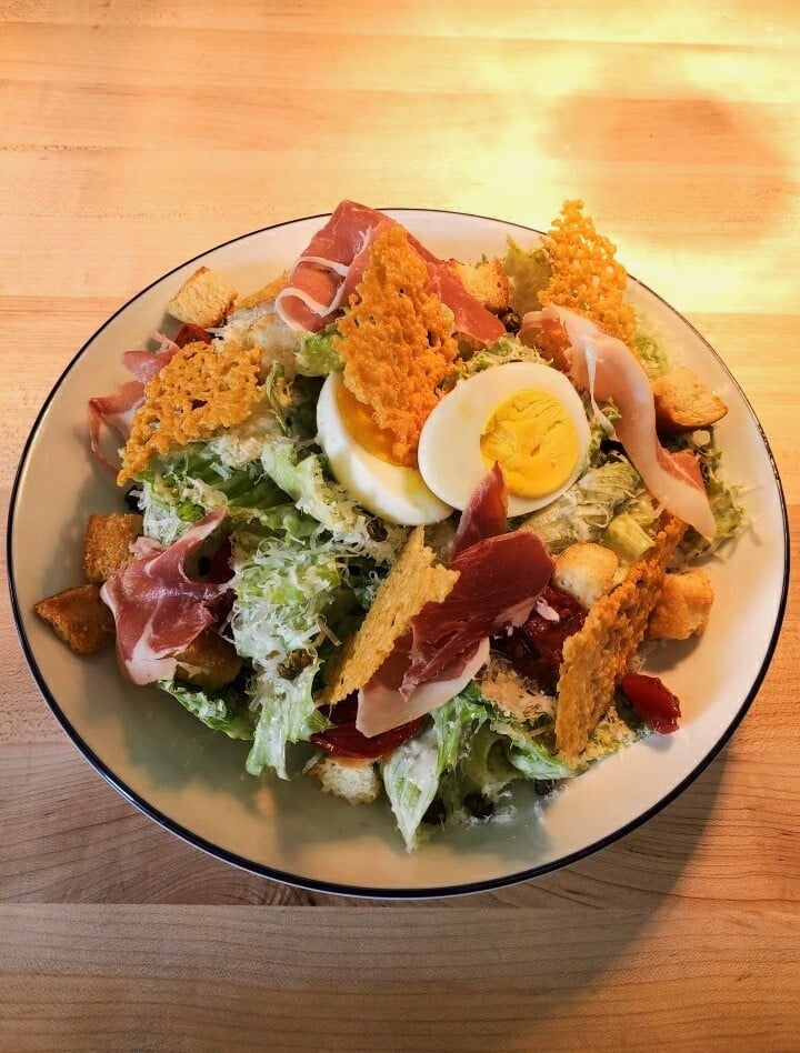 Image depicts a Caesar salad topped with crispy parmesan, boiled eggs and prosciutto.